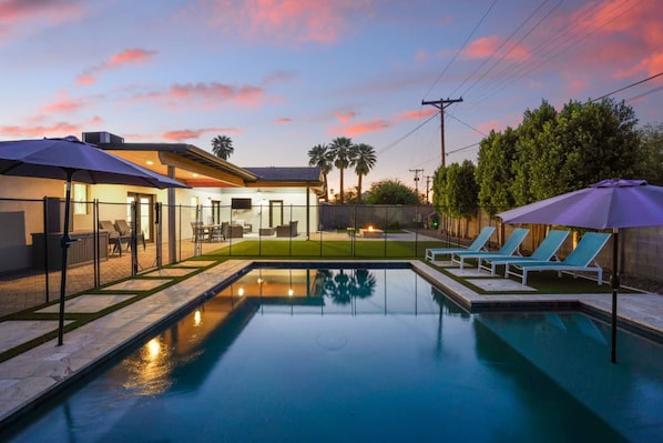 A swimming pool with lounge chairs and umbrellas, surrounded by a fenced patio area with outdoor furniture.