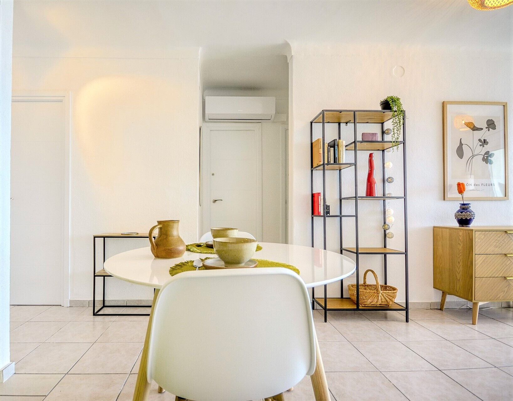 Et moderne spiseområde med et rundt bord, hvide stole, en reol og et skab.