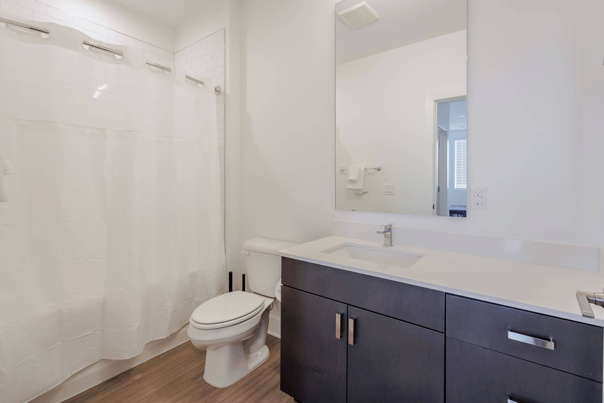 A modern bathroom with a white toilet, a shower with a white curtain, a large mirror, and dark cabinets.