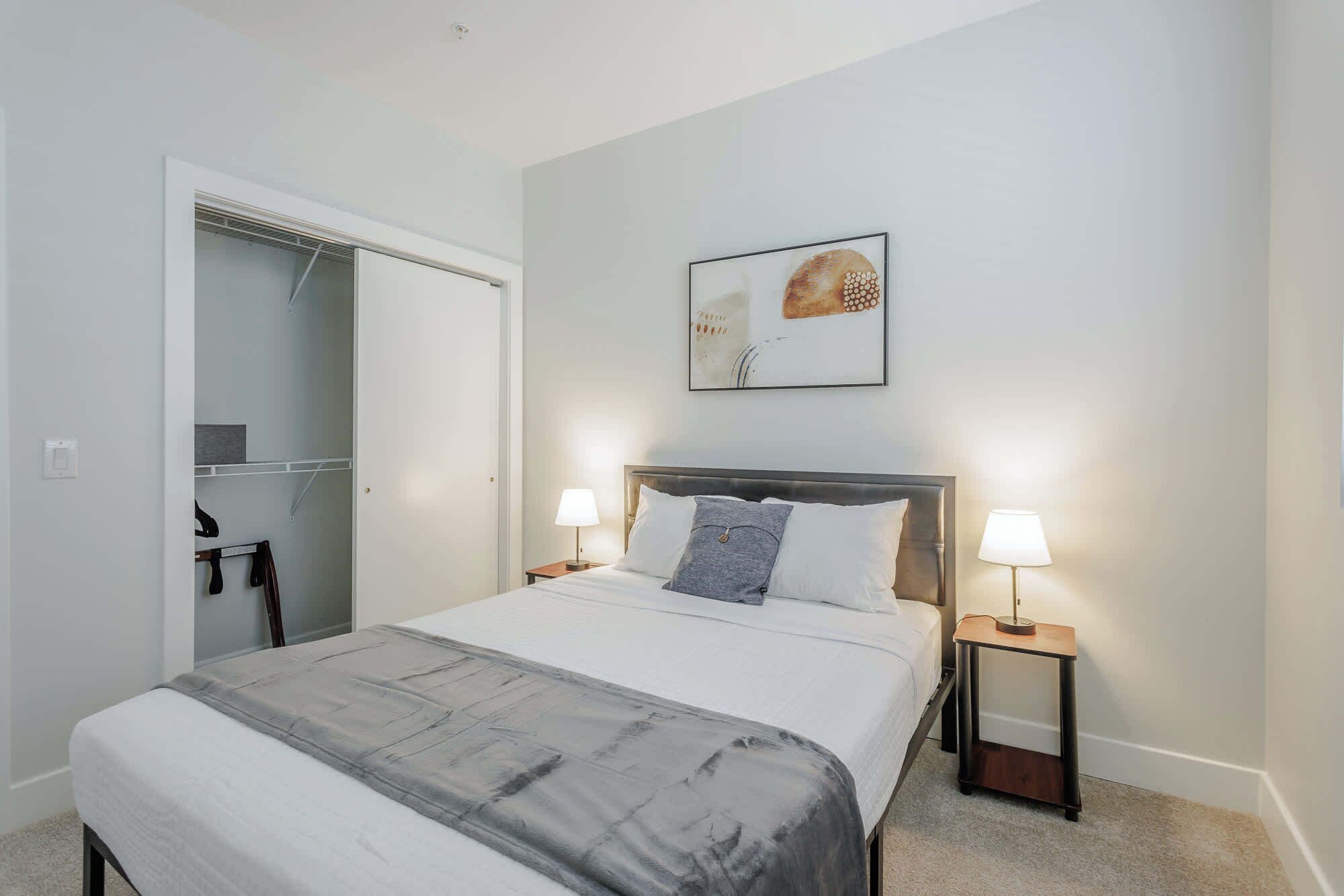 A bedroom with a bed, two bedside tables with lamps, a wardrobe, and a wall-mounted artwork.