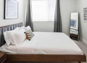 Una cama bien hecha con sábanas blancas y un cojín estampado, una mesita de noche, un espejo de pie y una ventana con cortinas.