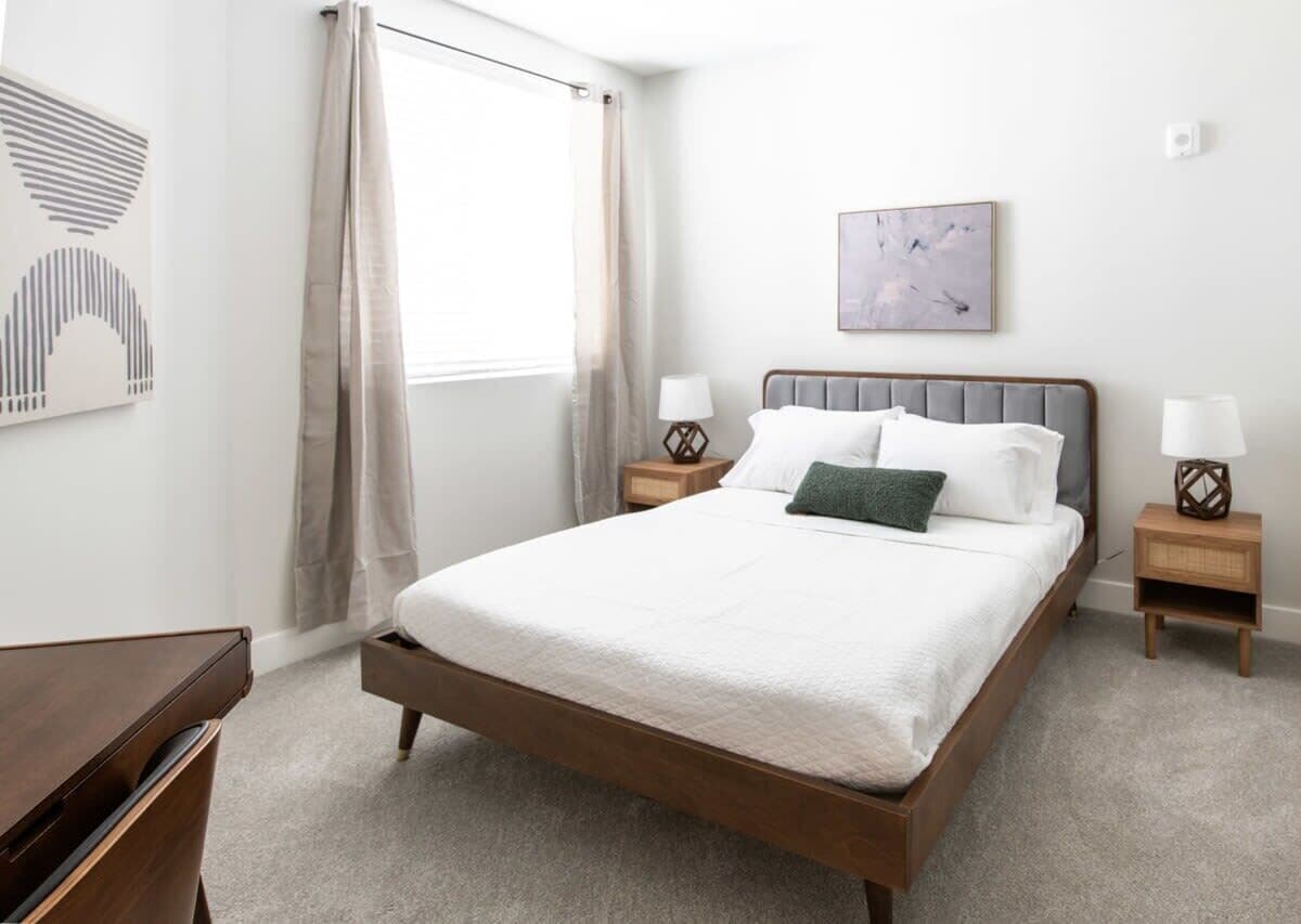 Une chambre à coucher bien aménagée, avec un grand lit, deux tables de chevet, une fenêtre avec des rideaux et une œuvre d’art accrochée au mur.