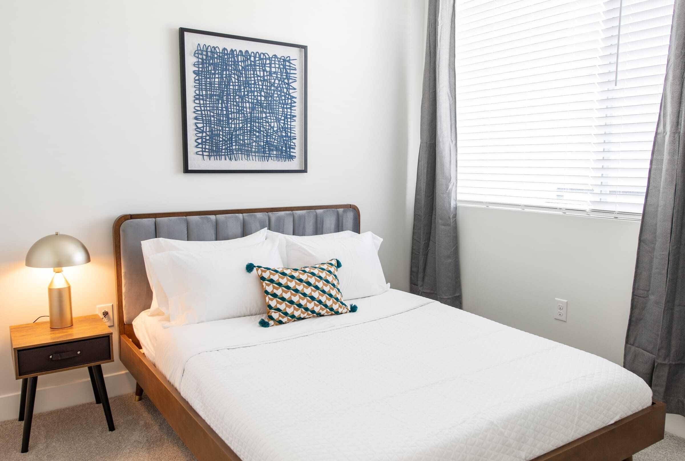 A neatly made bed with white linens and a patterned pillow, a bedside lamp, and a framed abstract artwork on the wall.