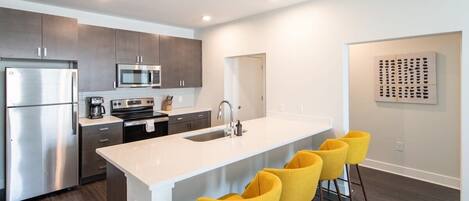 A modern kitchen with stainless steel appliances, a white island with bar stools, and a minimalist wall art piece.