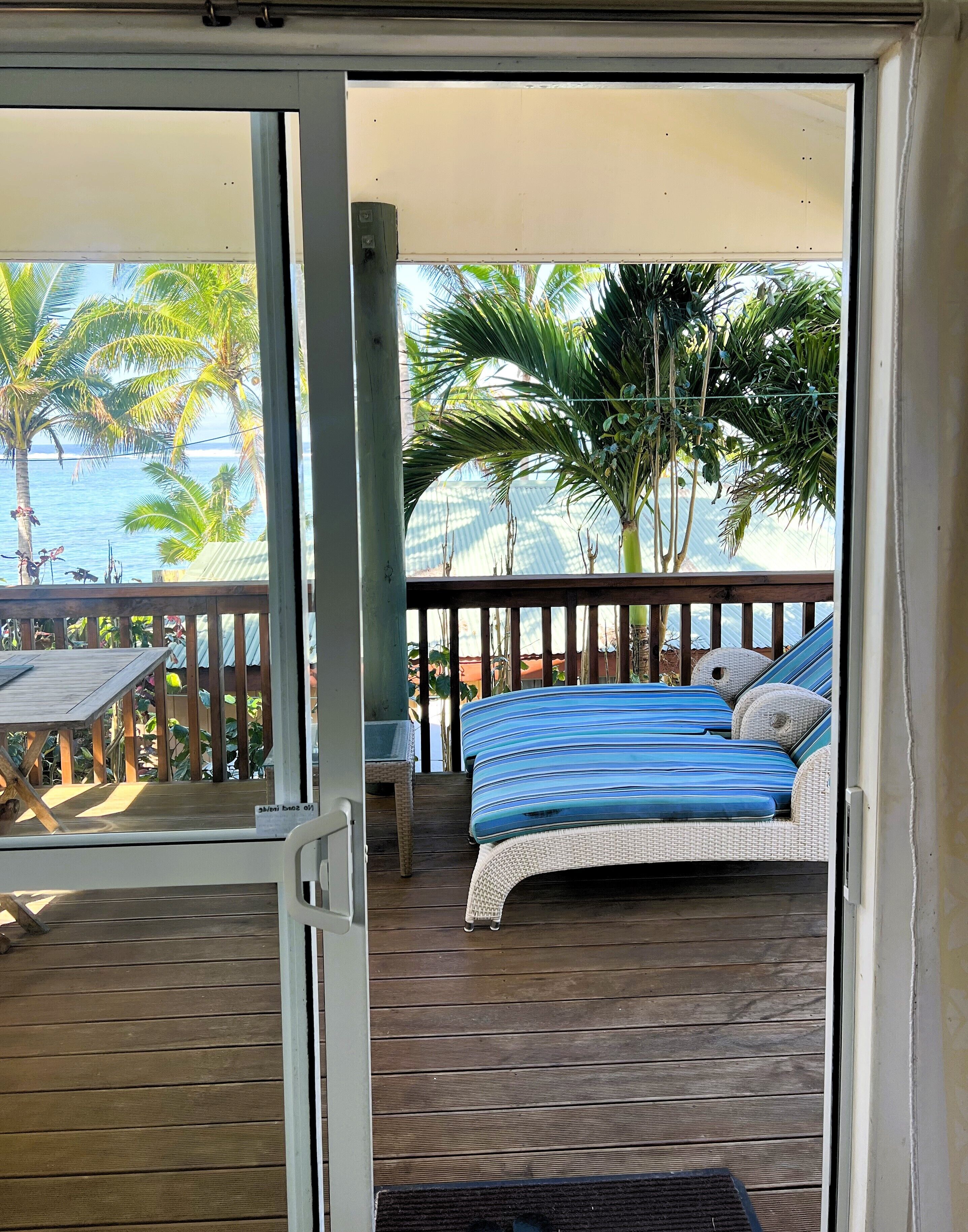 A balcony with a bed, a table, and a view of the ocean and palm trees.
