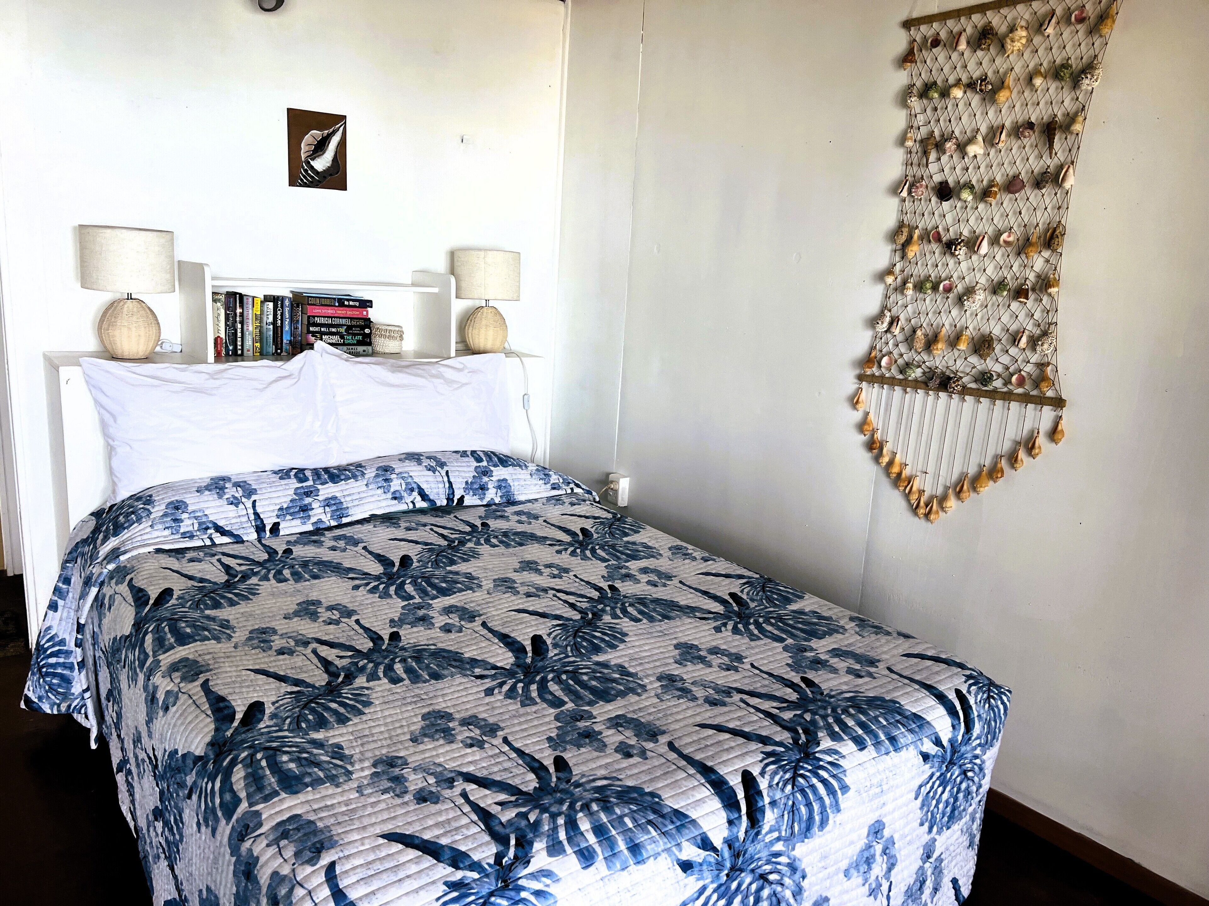 A neatly made bed with a floral-patterned bedspread, a bedside table with books and a lamp, and a wall hanging with various decorative items.