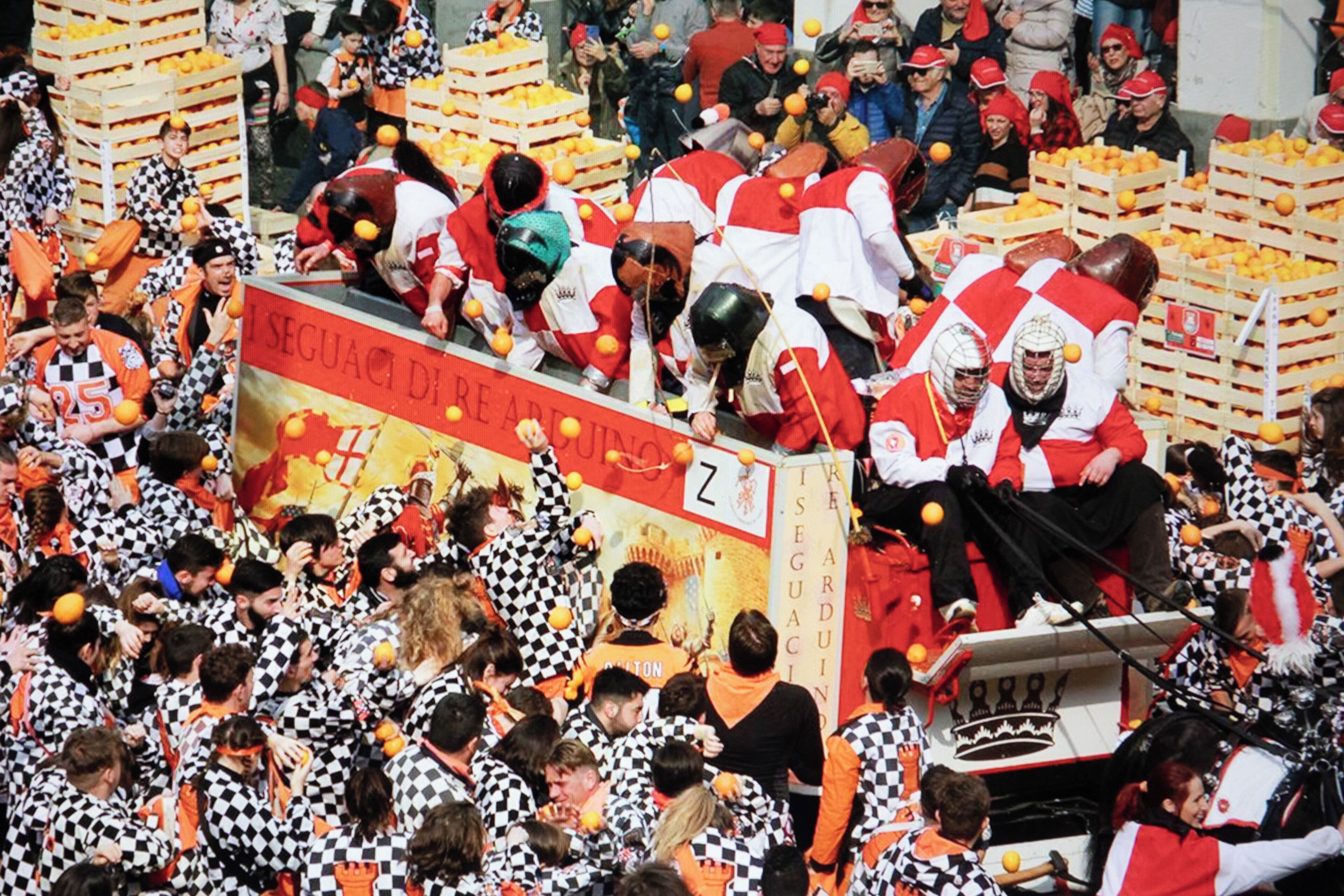 Un evento concurrido con participantes en uniformes a cuadros y detalles naranjas, rodeado de cajas de naranjas.