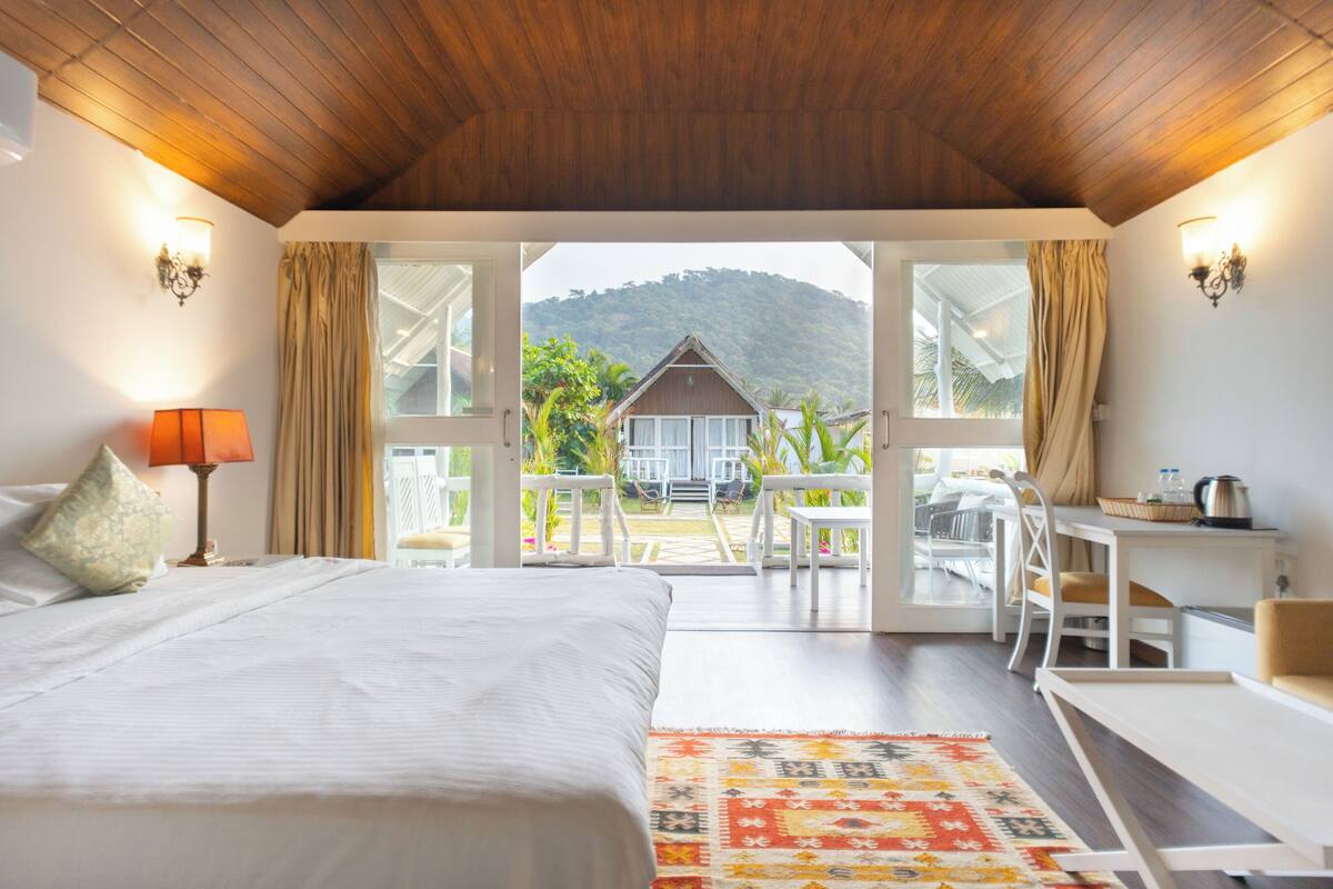 A bedroom with a large bed, a desk, and a view of a house and mountains through open doors.