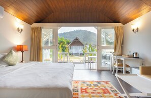 A bedroom with a large bed, a desk, and a view of a house and mountains through open doors. - Royal Agonda (Canacona)