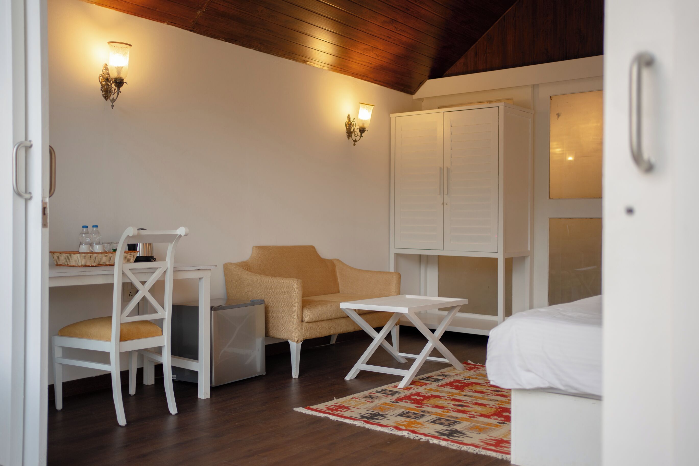 Una habitación con un escritorio blanco, un sofá color beige, una mesita blanca y una cama con ropa de cama blanca. Hay dos luces fijadas a la pared y un armario blanco.