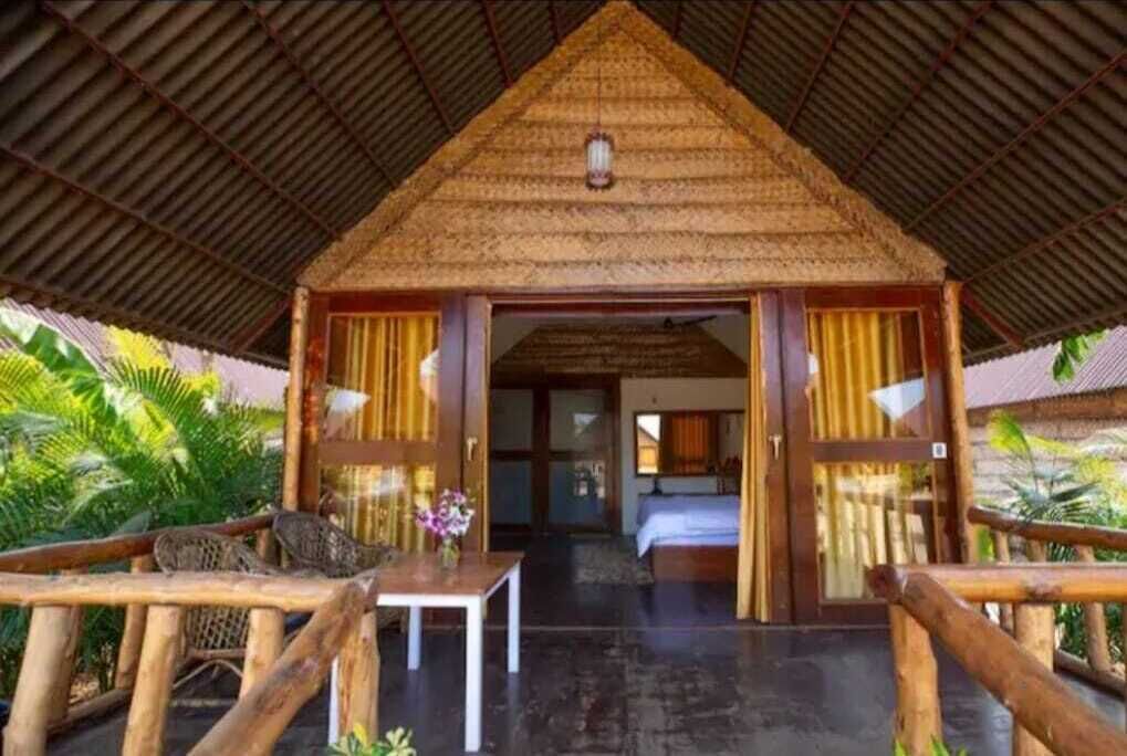 Een rieten hut met een bed en een tafel, omringd door houtenleuningen en groen.