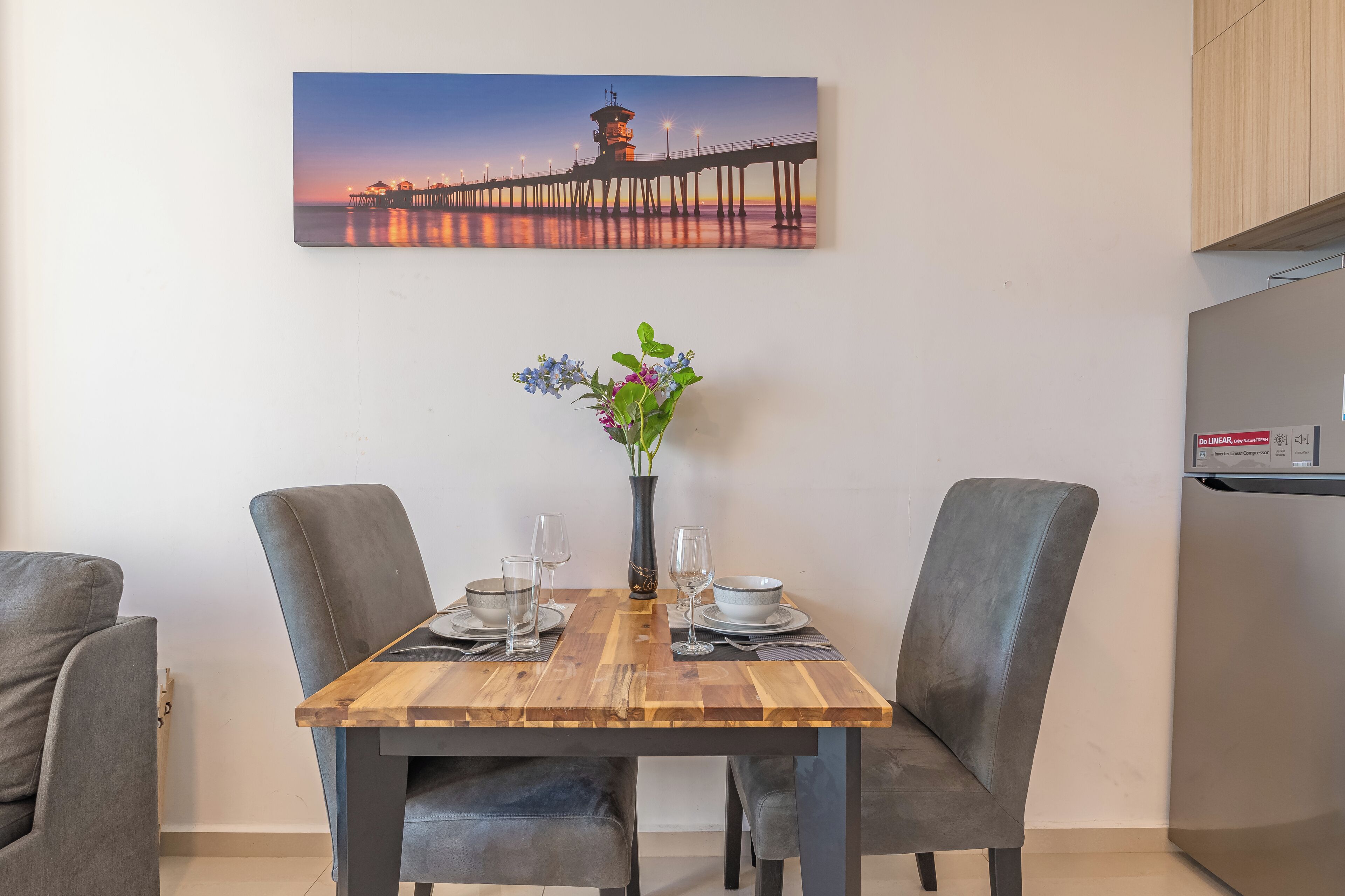 A dining area with a wooden table, two grey upholstered chairs, a vase with flowers, and a painting of a pier on the wall.