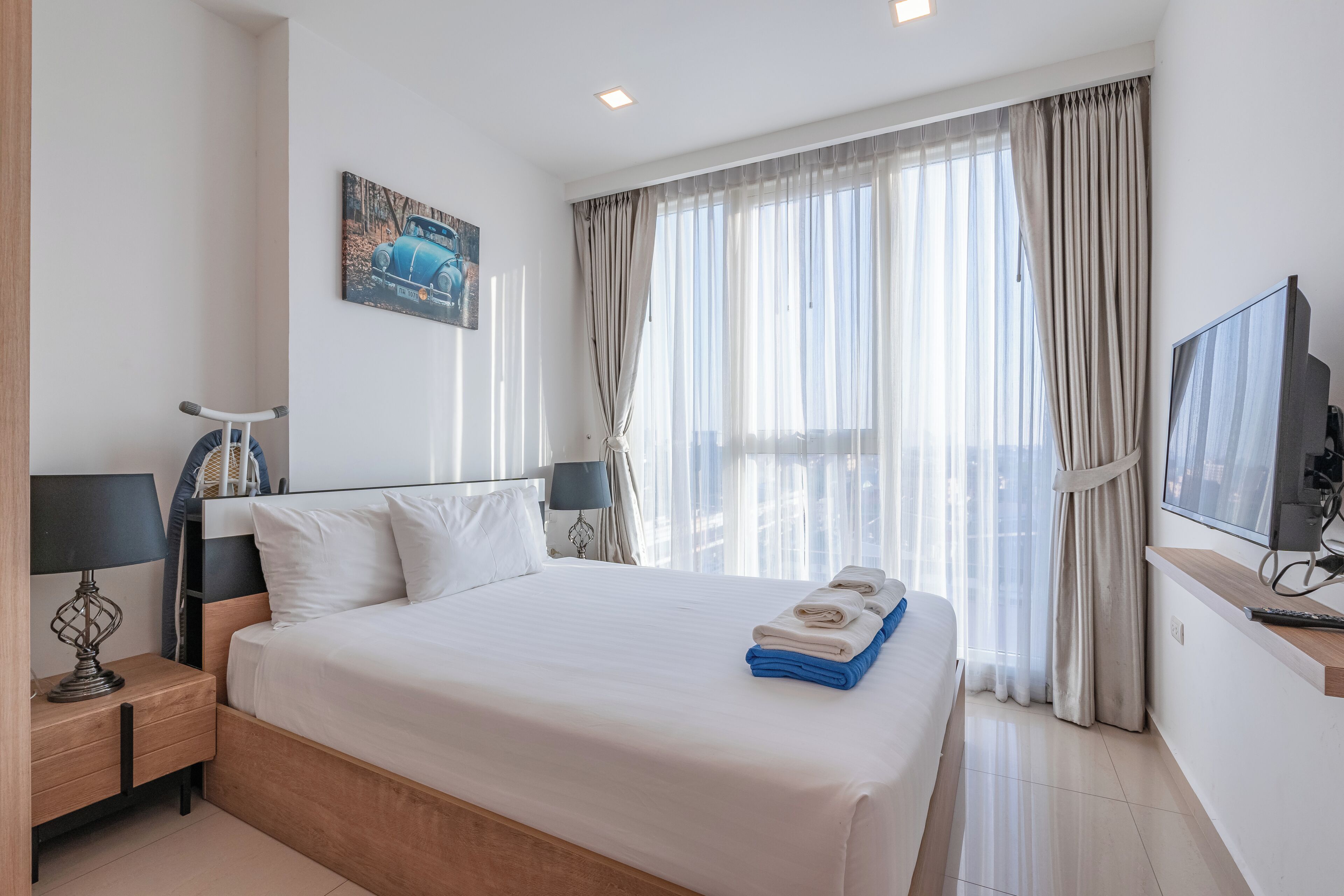 A hotel room with a large bed, a TV, a painting, and a window with curtains.