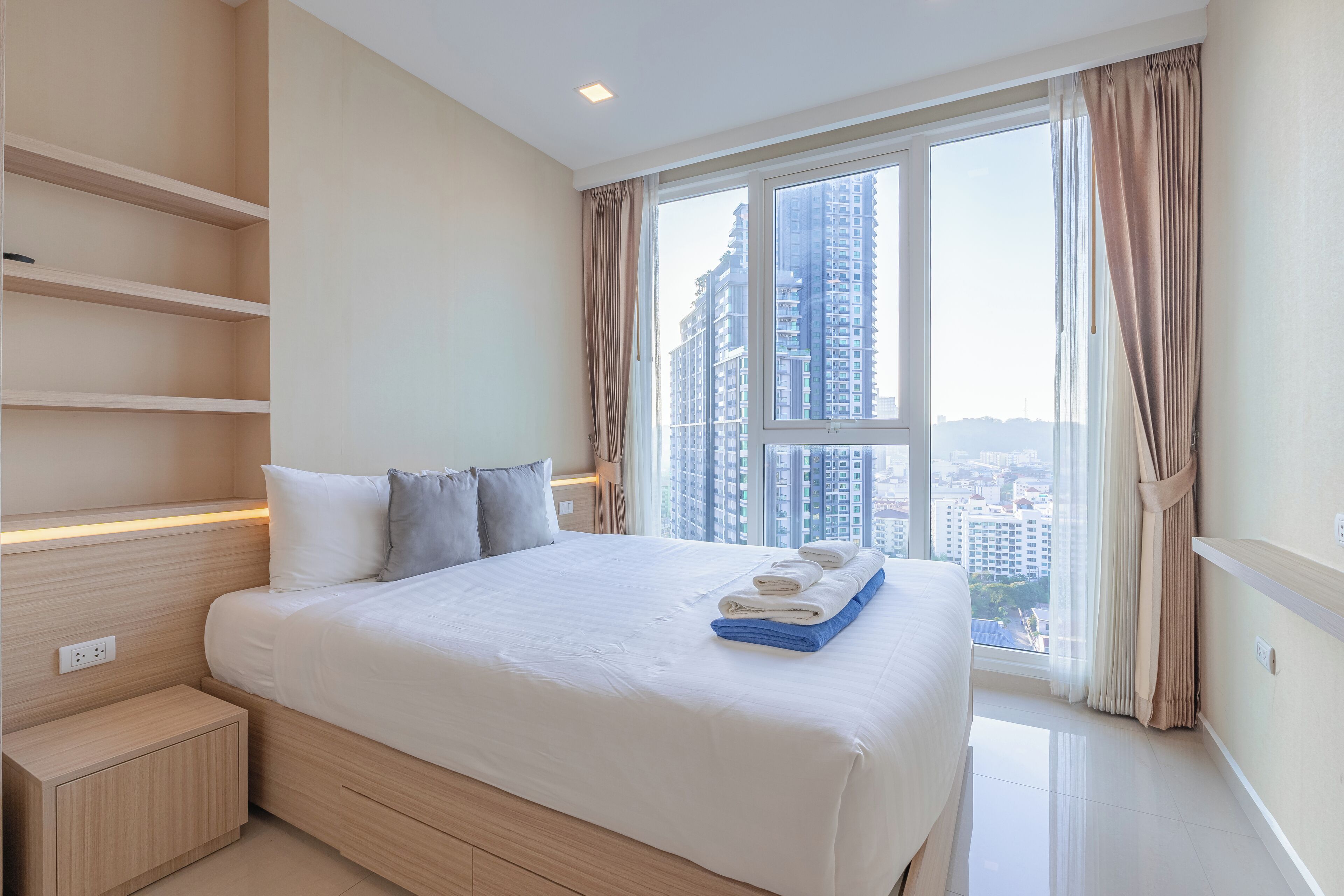 A modern hotel room with a large bed, built-in shelving, a view of tall buildings, and a window with curtains.
