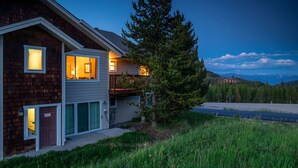 Une maison à deux étages avec une terrasse, un grand arbre à l’avant et une vue sur les montagnes en arrière-plan.