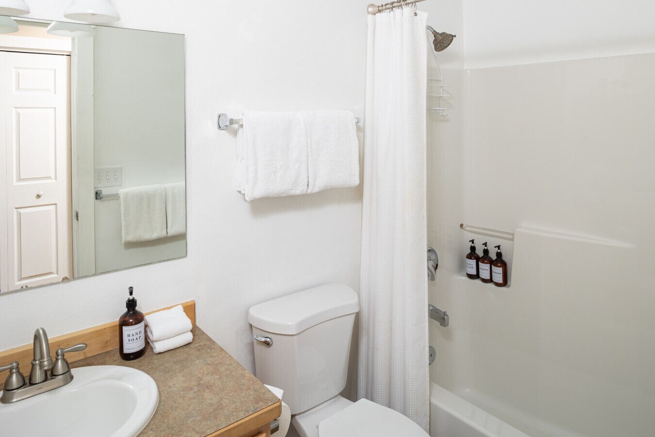 Un baño con lavamanos, inodoro y ducha. Hay botellas de jabón de manos y champú en la pared.