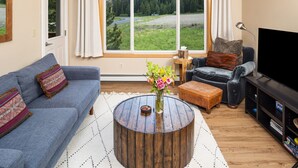 Un salon avec un canapé bleu, un fauteuil foncé, une table basse ronde ornée d’un vase de fleurs et un téléviseur posé sur un meuble noir.