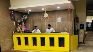 A reception area with a yellow desk, two staff members, a clock, and a sign reading "RECEPTION". - Hotel Ashoka International (New Delhi)