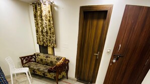 A room with a patterned sofa, a white chair, and a wooden door. - Hotel Ashoka International (New Delhi)