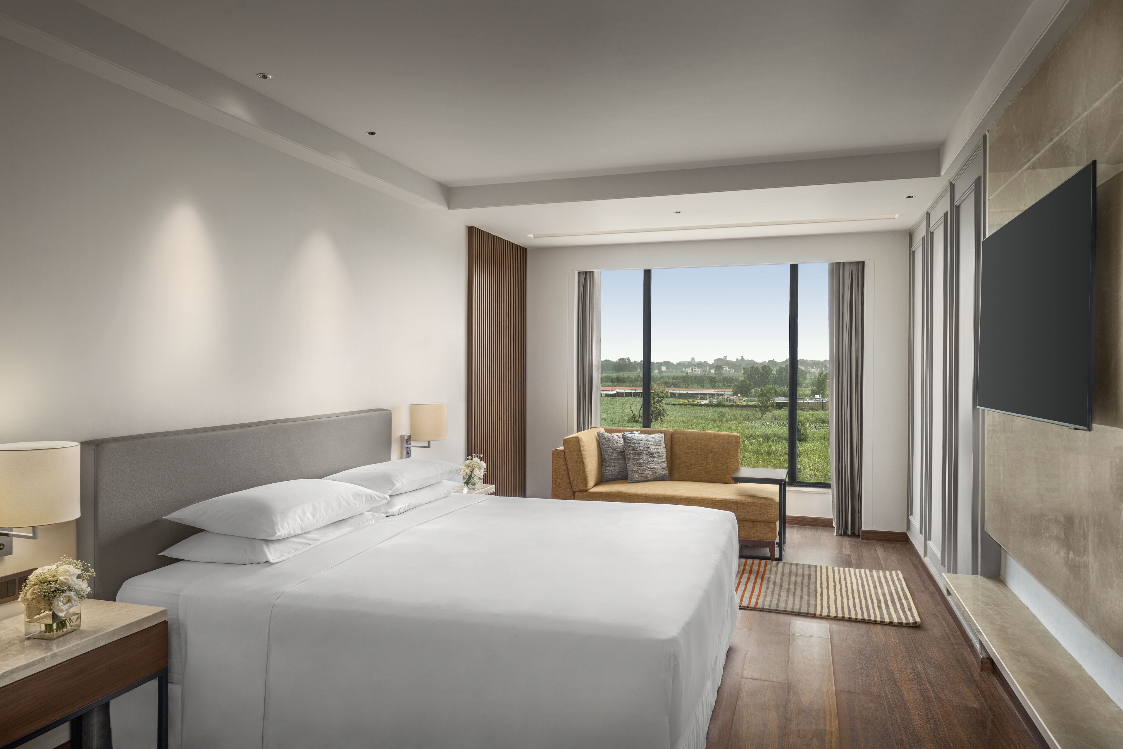 A modern hotel room with a large bed, a sofa, a TV, and a view of the city.