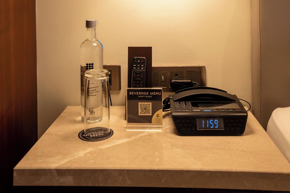 Ein Hotelzimmer mit Nachttisch, der eine Lampe, eine Minibar, eine digitale Uhr und eine Getränkekarte zeigt.