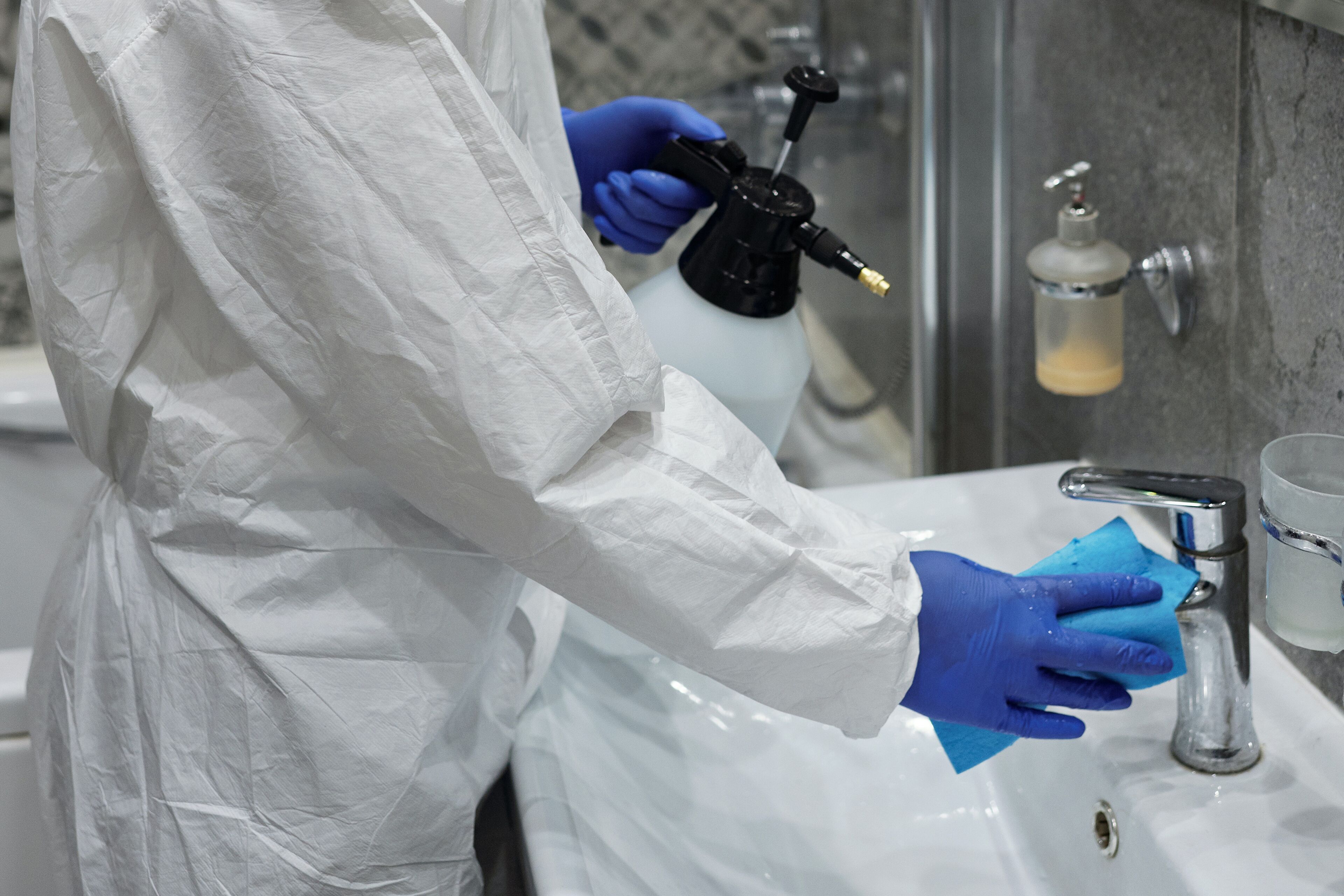A person in a white protective suit and blue gloves cleaning a sink with a spray bottle and a sponge.