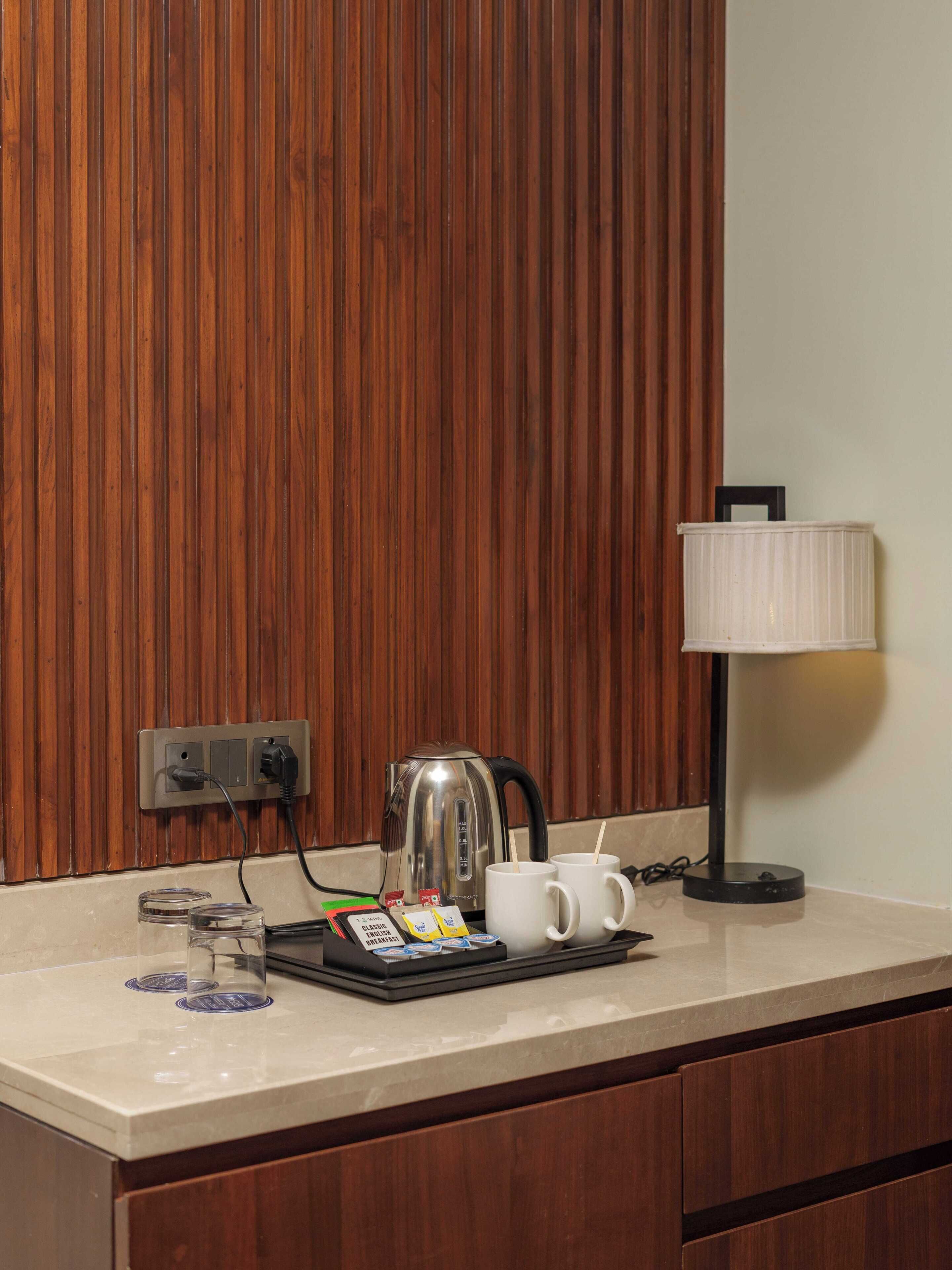 A kitchenette with a kettle, two mugs, a lamp, and a coffee maker on a countertop.