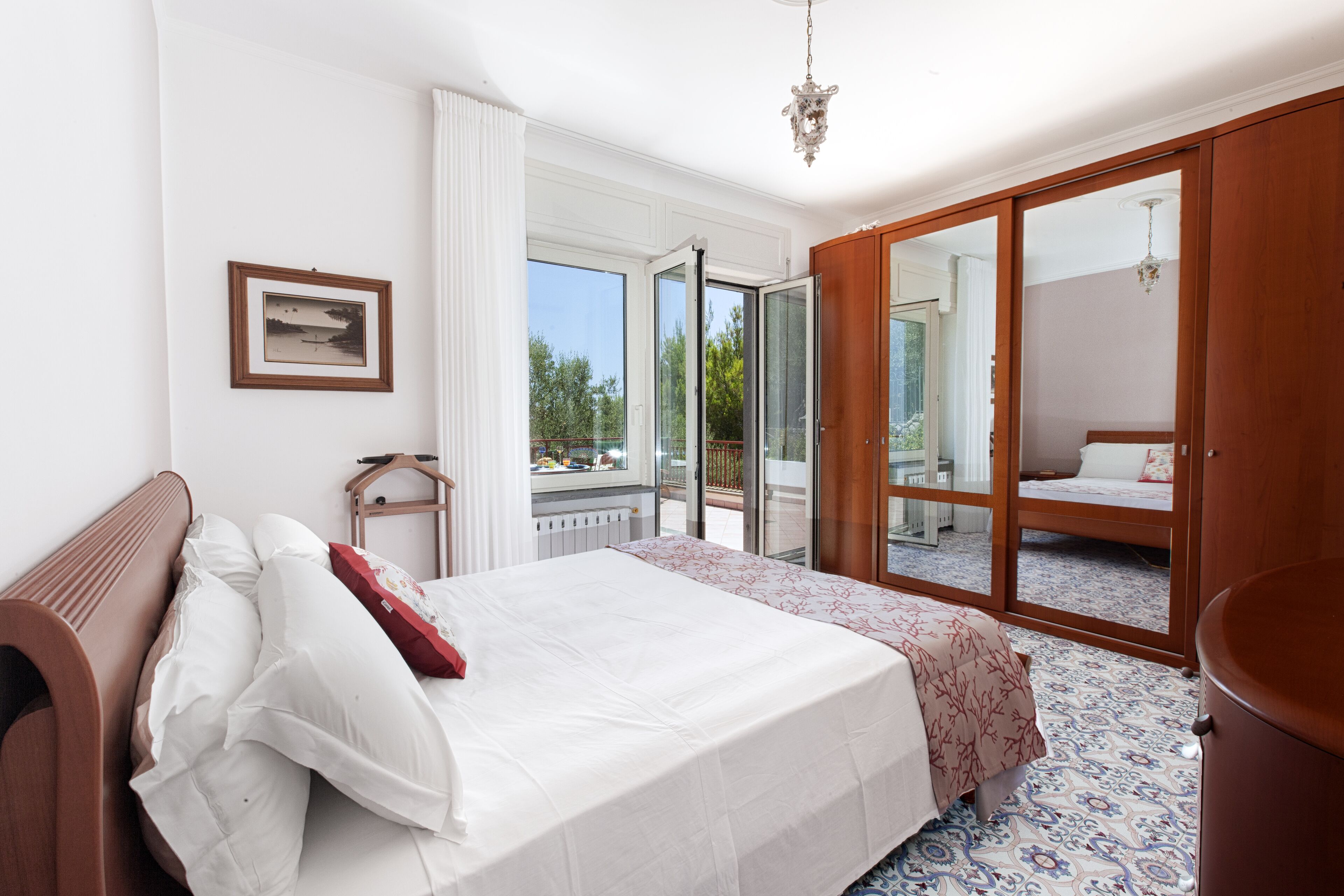 Ein Schlafzimmer mit einem großen Bett, einem Holzkopfstück, einem Fenster mit weißen Vorhängen, einem gerahmten Bild an der Wand und einem Balkon mit Blick auf Bäume.