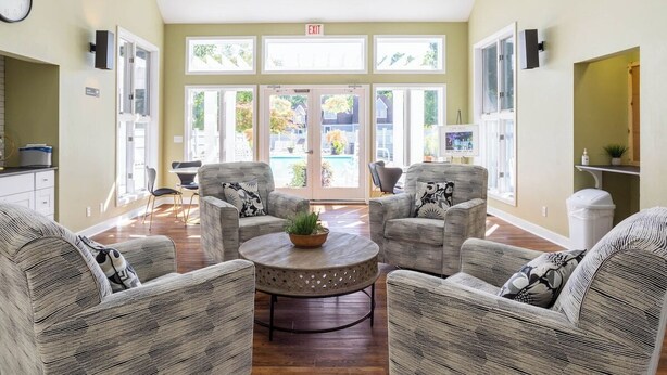A spacious lobby with a central round table, four grey upholstered chairs, and a large window offering a view of the outside.