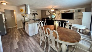 A modern kitchen with a central island, white cabinetry, and a dining area with a wooden table and white chairs. - Nearby North Dallas - Grandscapes (The Colony)