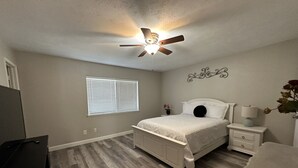 A bedroom with a bed, nightstands, a ceiling fan, a window with blinds, and a wooden floor. - Nearby North Dallas - Grandscapes (The Colony)