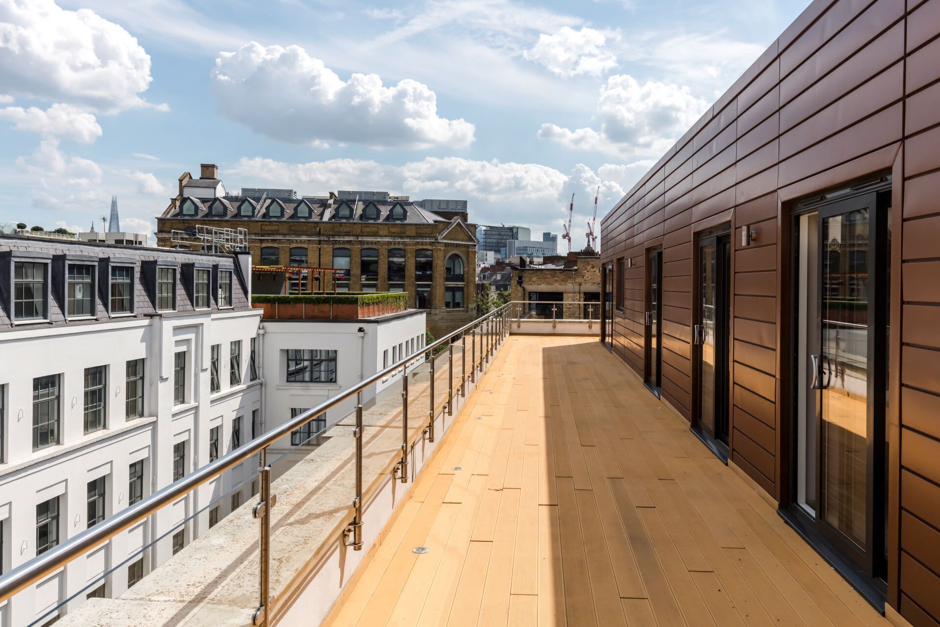 Eine Dachterrasse mit Holzboden, Metallgeländern und großen Fenstern, die einen Blick auf umliegende Gebäude und den Himmel bietet.