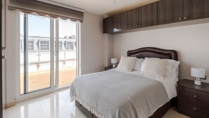 Quarto com uma grande porta de vidro deslizante que dá para uma varanda, cama com cabeceira, duas mesas de cabeceira com abajures e um guarda-roupa de madeira acima da cama.