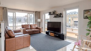 Uma sala de estar moderna com um sofá de couro, uma televisão de tela plana, uma poltrona de balanço de madeira e uma varanda com vista para edifícios.