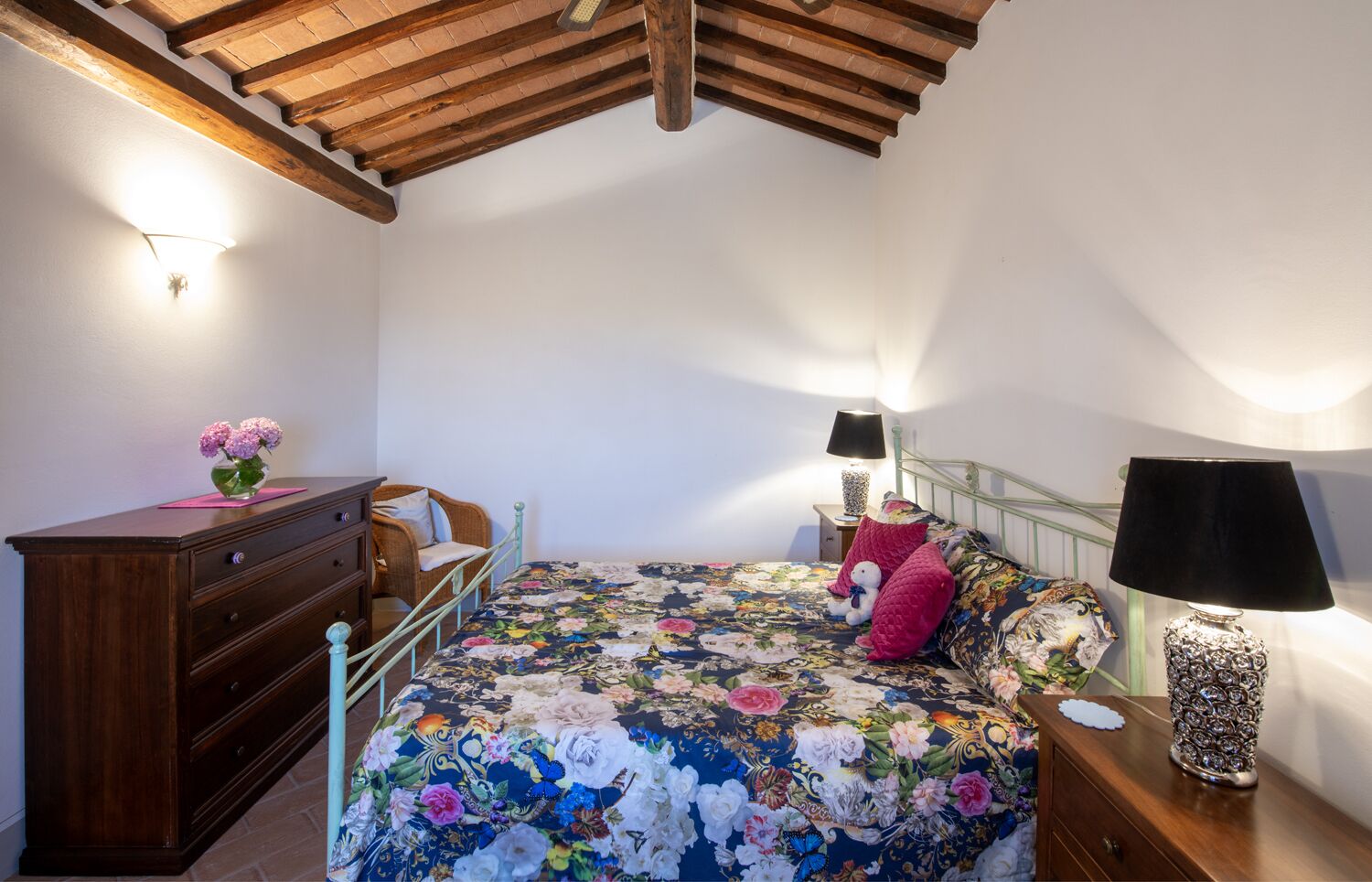 Une chambre avec un plafond en bois, une couvre-lit à motifs floraux, une commode en bois, une chaise, deux lampes de chevet et un vase décoratif.