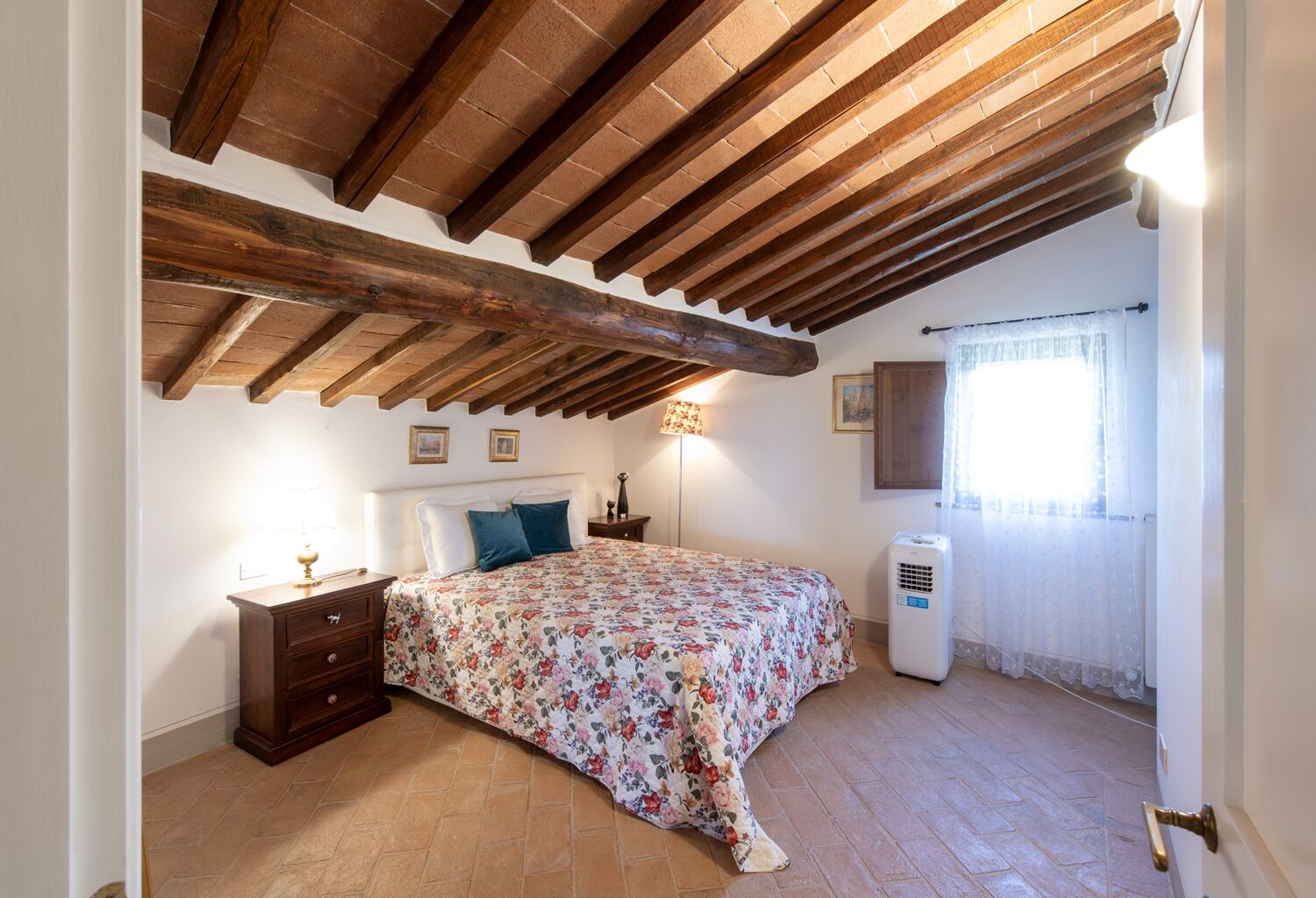 A bedroom with a wooden ceiling, a bed with floral bedding, a nightstand, a lamp, and a window with curtains.