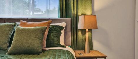 A neatly made bed with green bedding, a wooden nightstand with a lamp, and a view of trees through a window.
