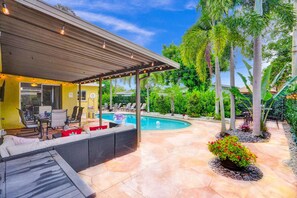 Un espacio de estar al aire libre cubierto con piscina, rodeado de plantas tropicales y una pérgola con luces colgantes.