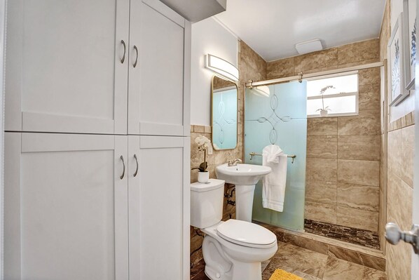 Baño con inodoro, lavabo y espejo. Cuenta con una zona de ducha con puerta de vidrio y un perchero para toallas.