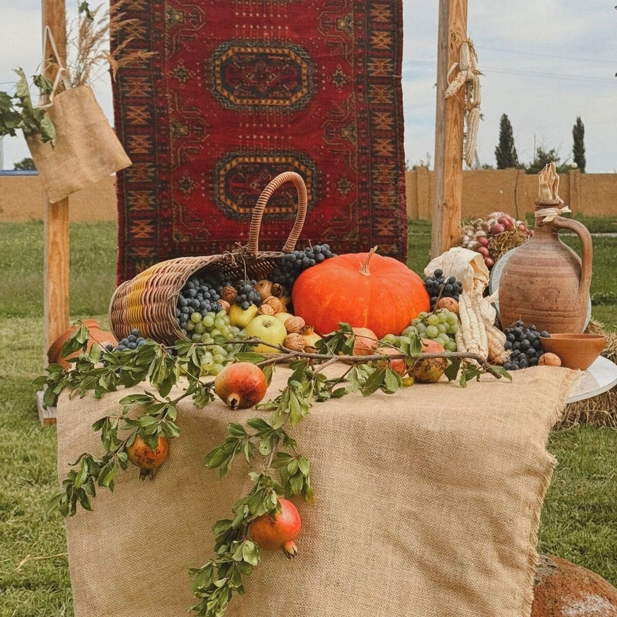 A table with a variety of fruits and vegetables, a woven basket, a ceramic jug, and a rustic setting with a patterned rug and wooden poles.