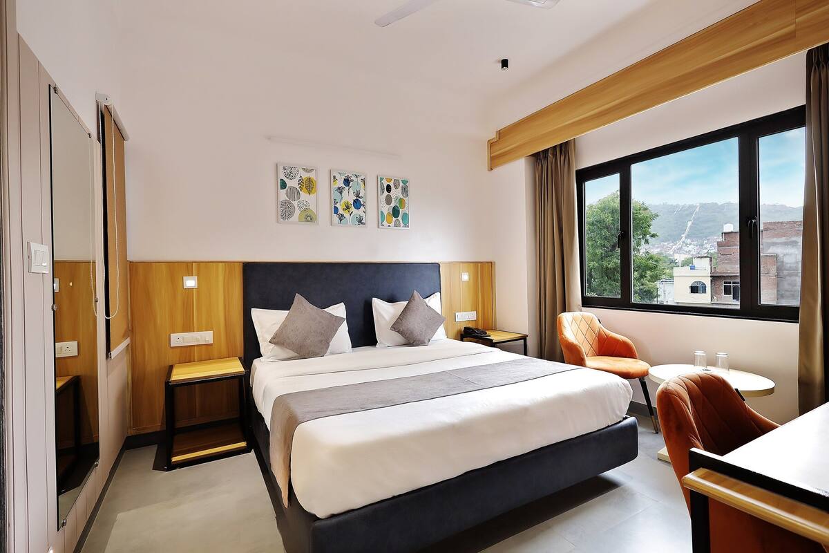 Habitación de hotel con una cama grande, un escritorio, una silla y un ventanal con vistas a edificios y árboles.
