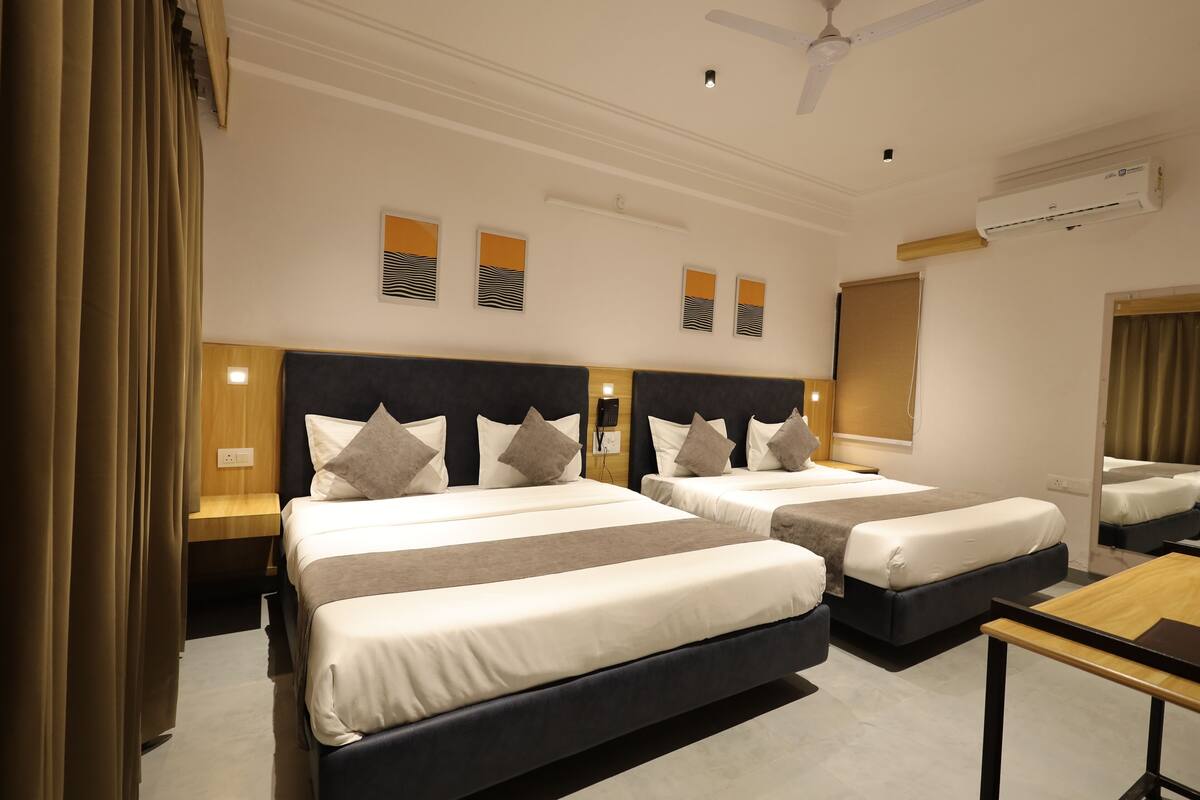Una habitación de hotel con dos camas, un ventilador de techo, un equipo de aire acondicionado y una mesita de noche de madera con lámpara.