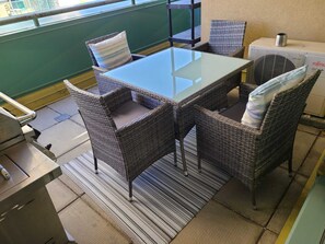 Een balkon met een glazen tafel, rieten stoelen, een barbecue en uitzicht over de stad.