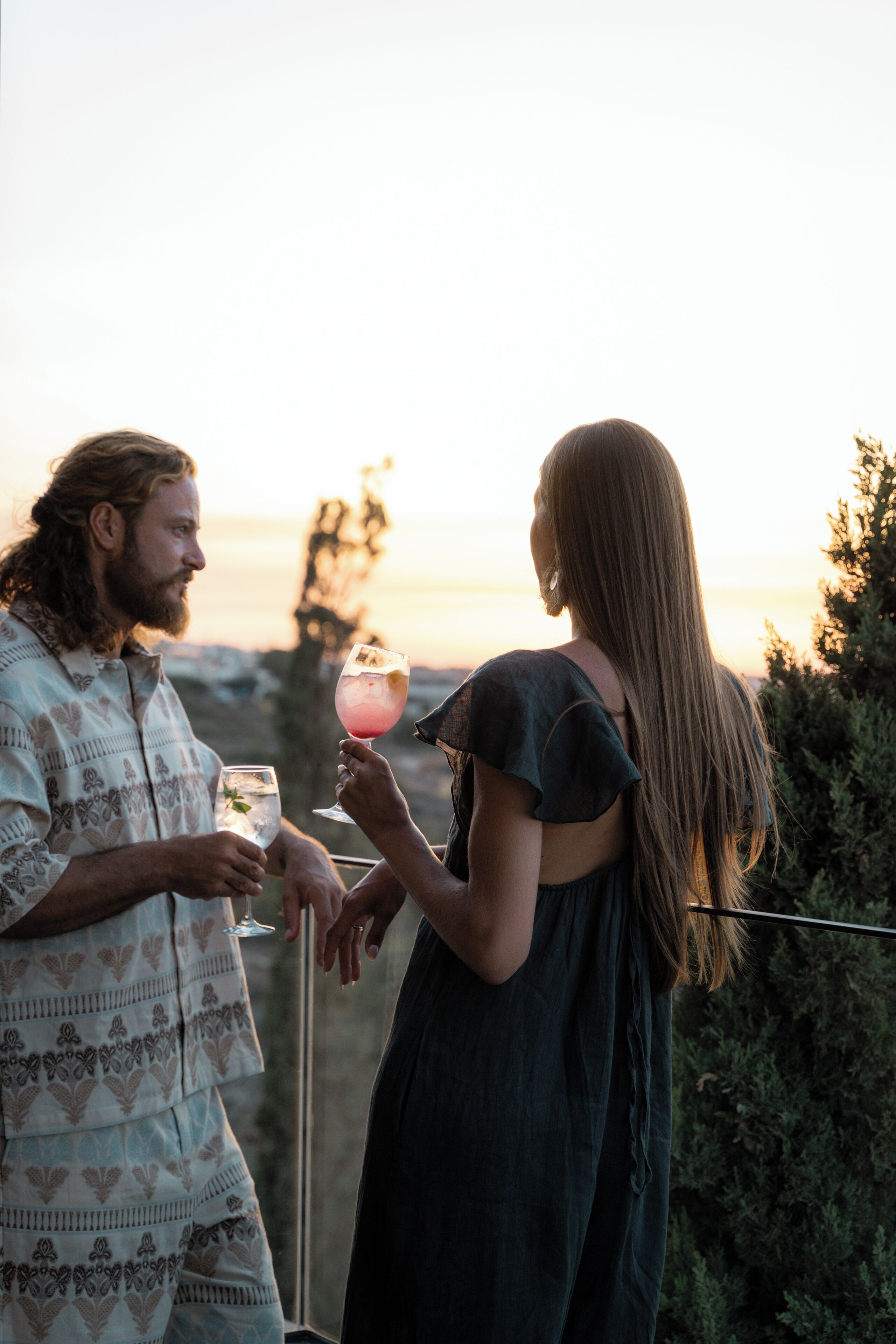 Two people holding drinks at an outdoor setting with a view of the sunset.