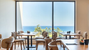 Un espace repas doté de grandes fenêtres donnant sur la mer, de tables et de chaises en bois, et offrant une vue sur l’océan.
