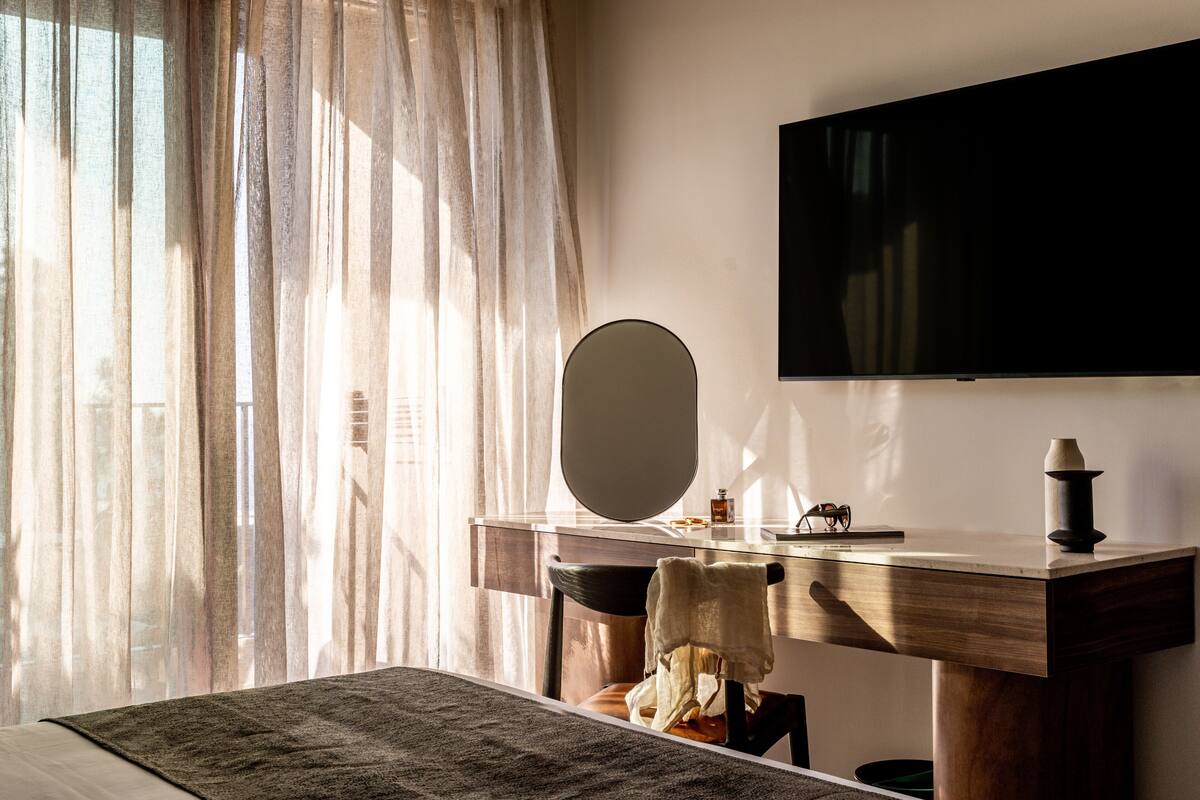 A bedroom with a bed, a bedside table, a mirror, a flat-screen TV mounted on the wall, and sheer curtains.
