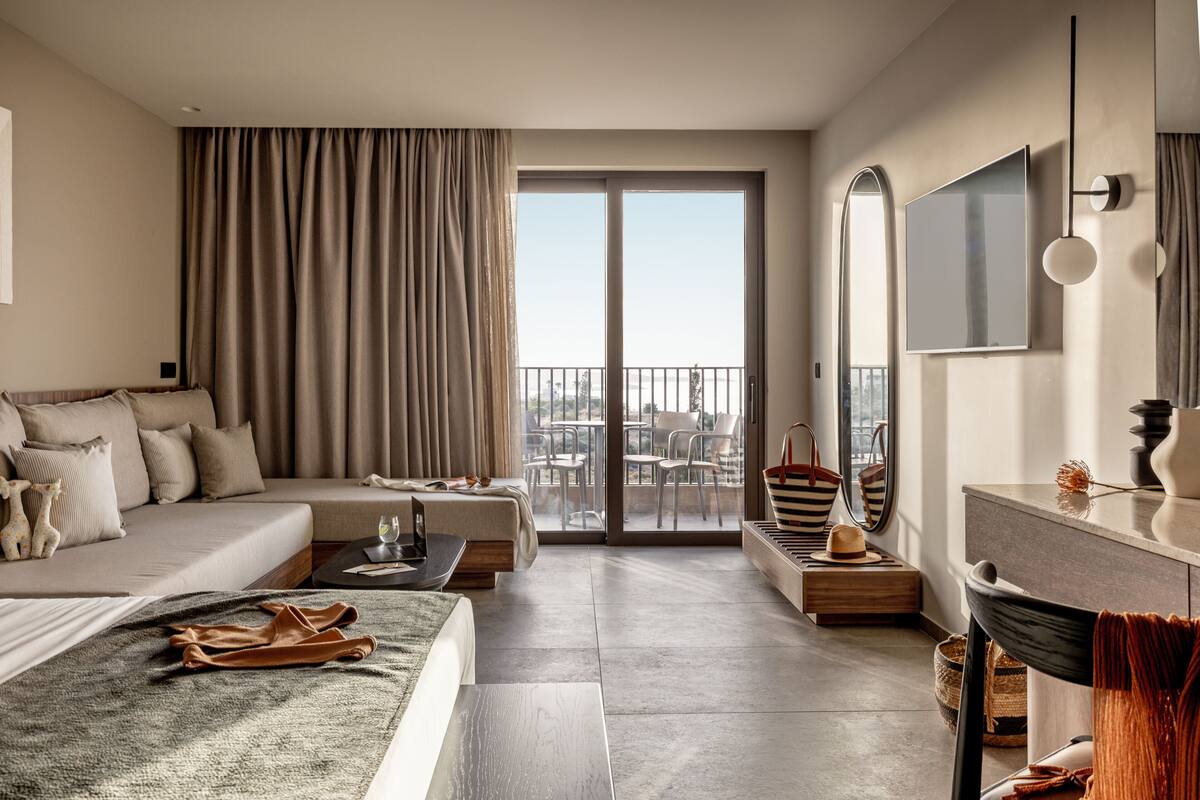 A modern hotel room with a large bed, a sofa, a desk, and a balcony with a city view.