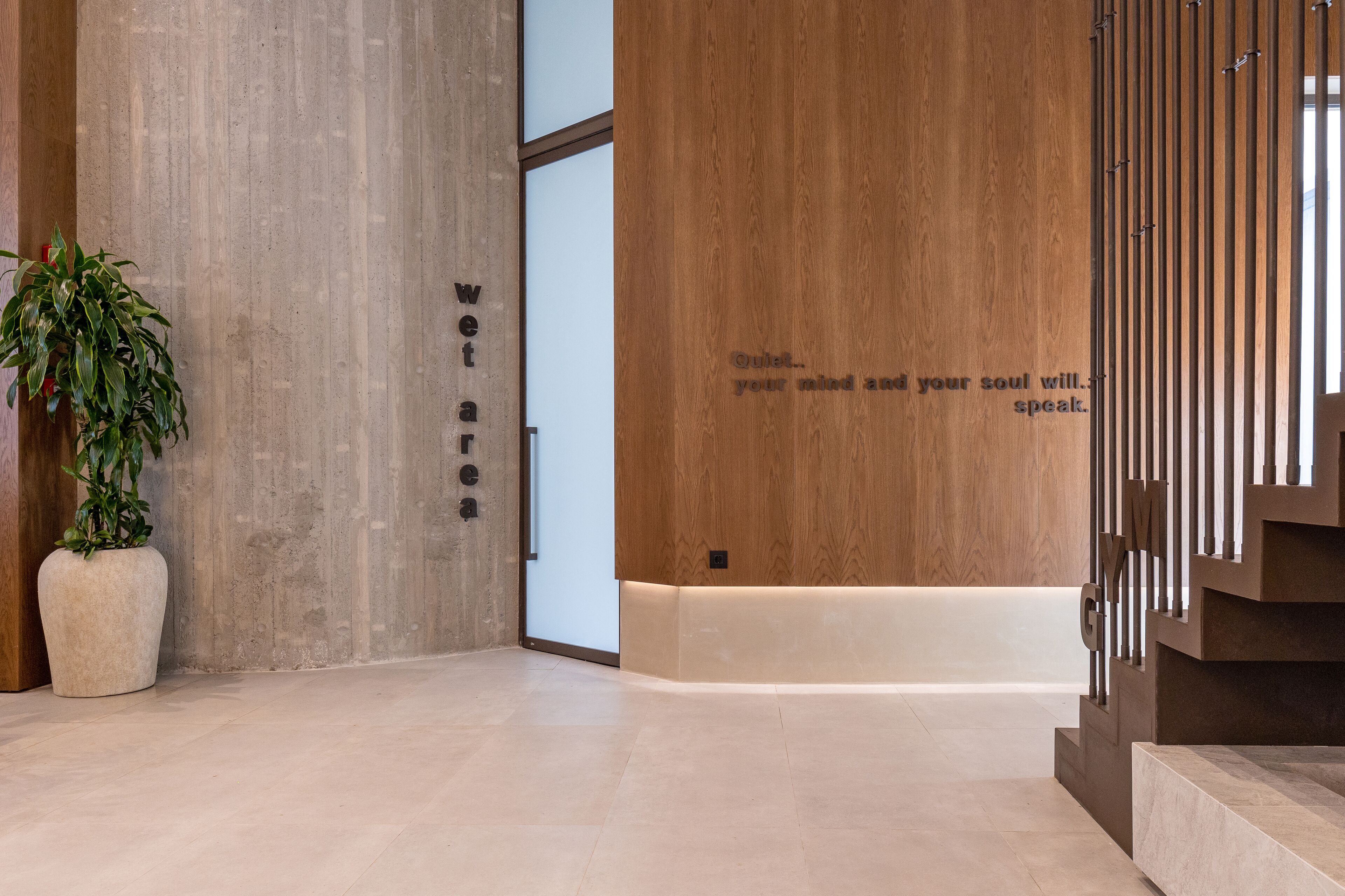 A modern lobby with a concrete wall, a wooden panel with a quote, a potted plant, and a staircase.
