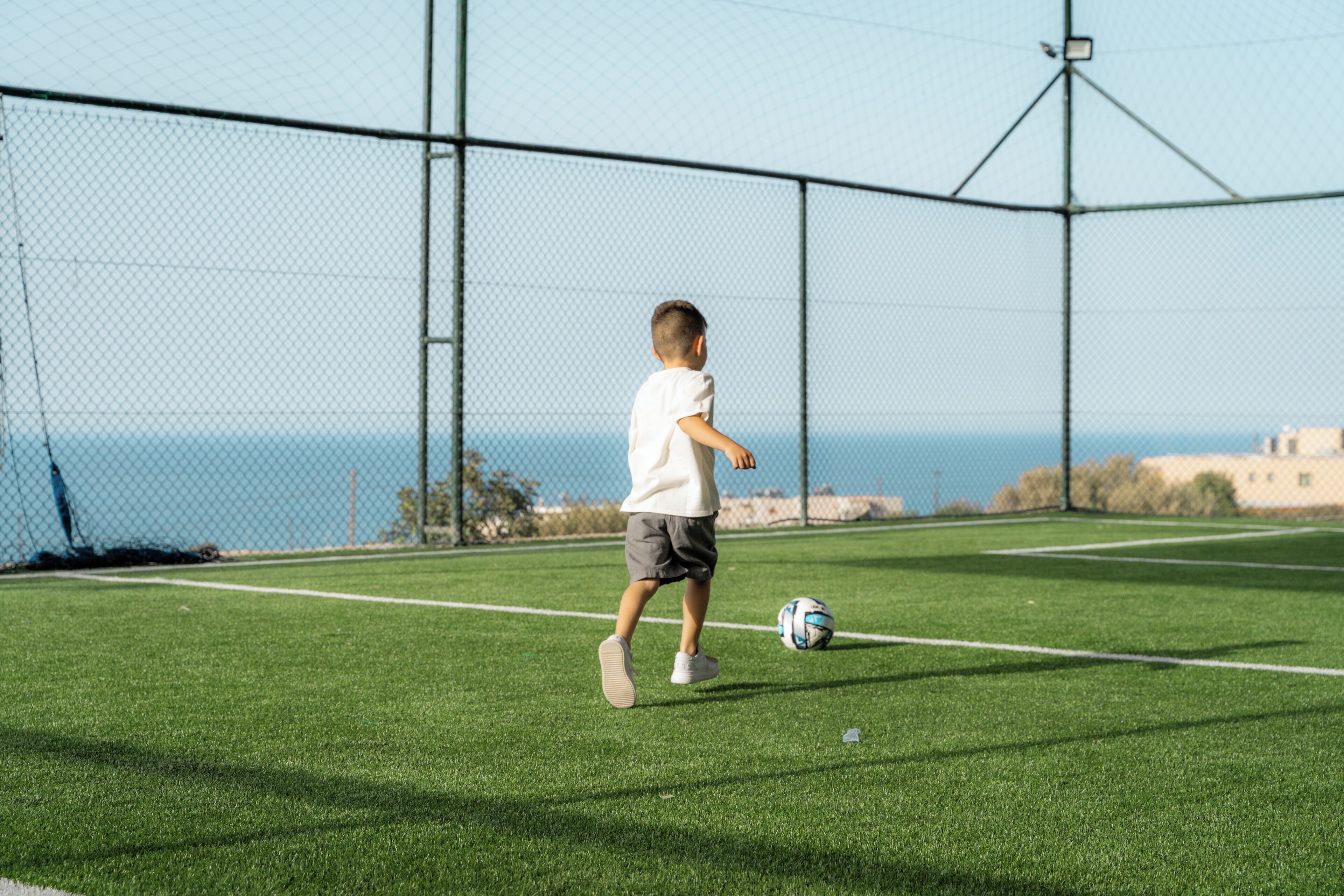 Un enfant joue au football sur un terrain verdoyant, avec une clôture et un ciel dégagé en arrière-plan.