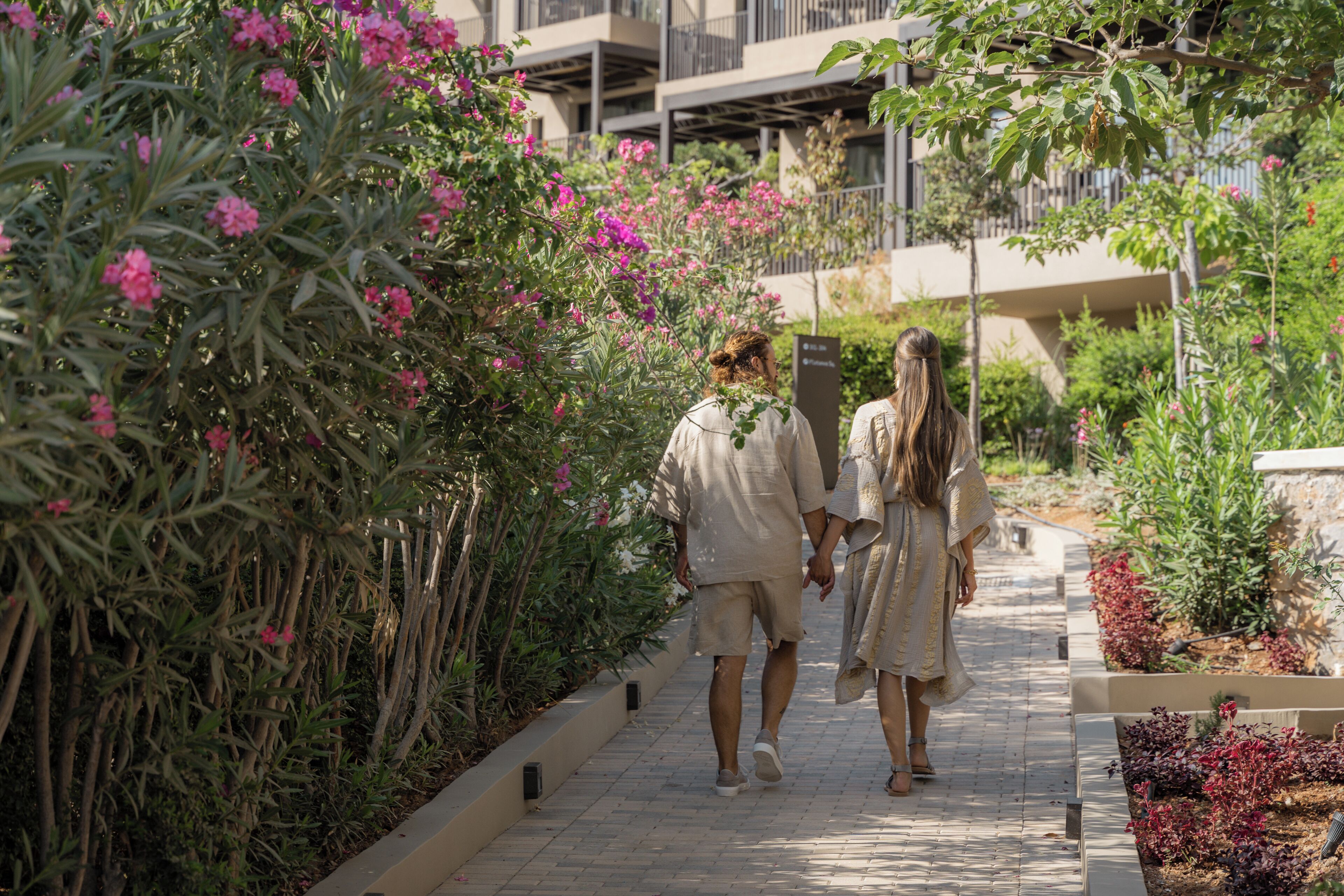 Un chemin bordé de fleurs roses et de verdure, menant à un bâtiment doté de balcons.